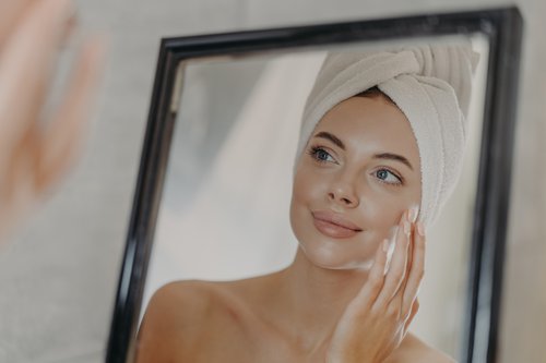 Young naked European woman touches soft skin on face, looks at herself in bathroom mirror, wears wrapped white towel on head after taking bath, poses bare shoulders. Beauty and skin care concept