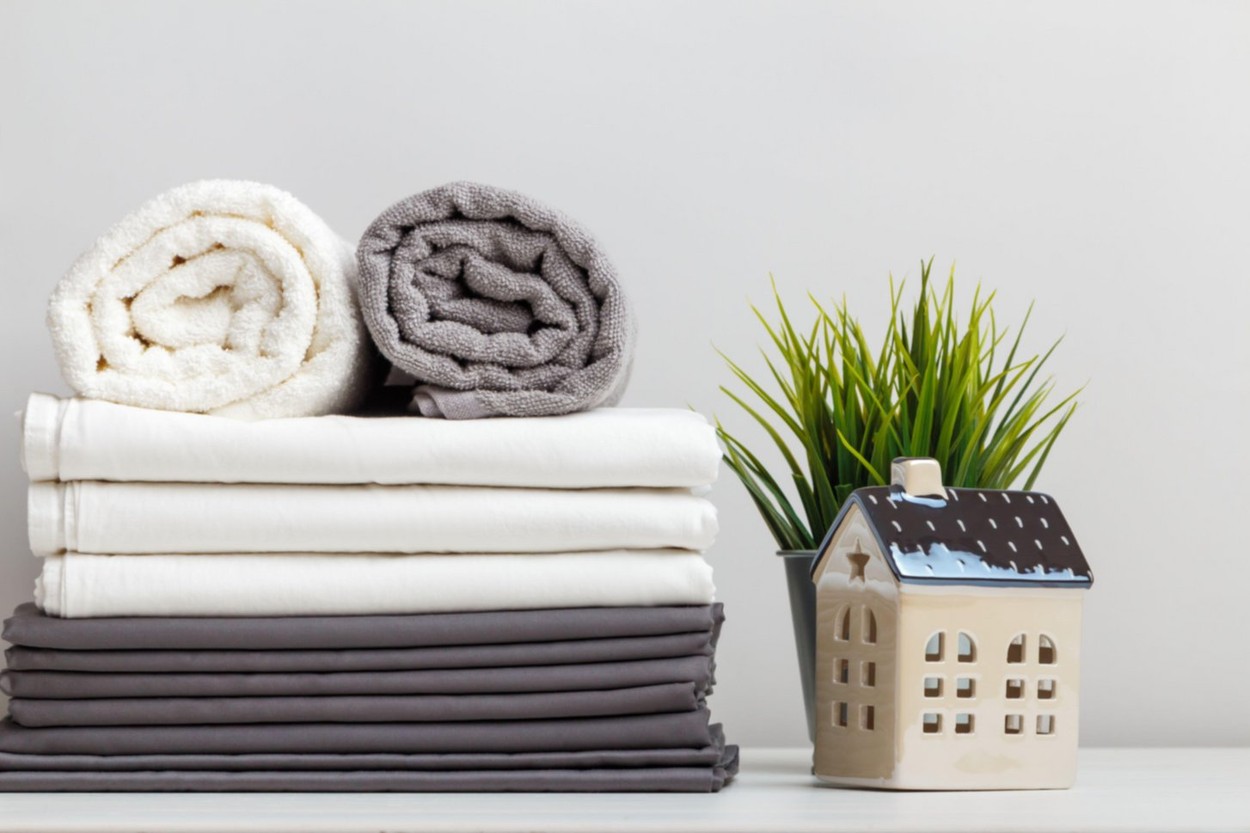 A stack of gray and white linens, sheets and terry towels on the table. A potted plant and a toy little house