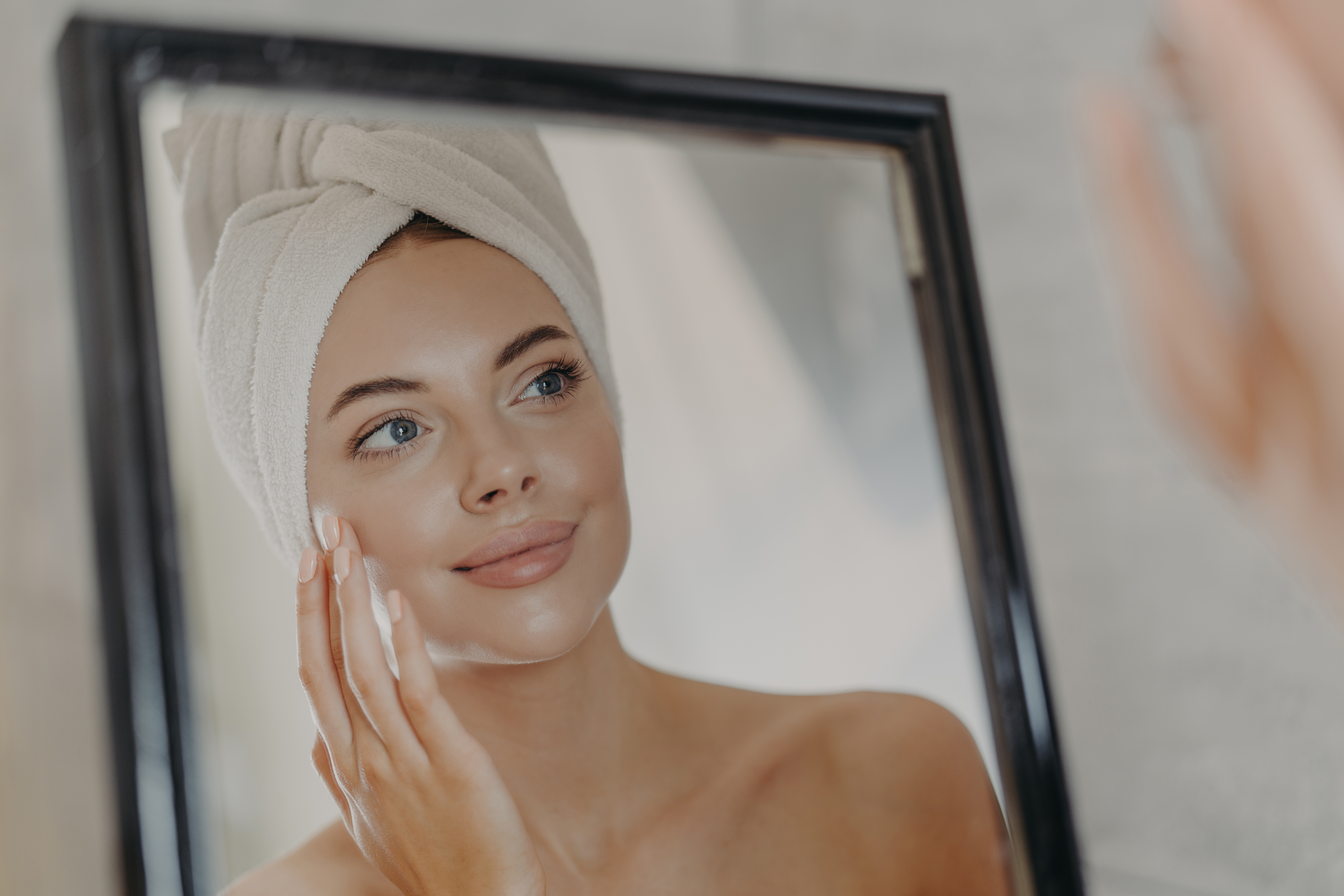 Young naked European woman touches soft skin on face, looks at herself in bathroom mirror, wears wrapped white towel on head after taking bath, poses bare shoulders. Beauty and skin care concept