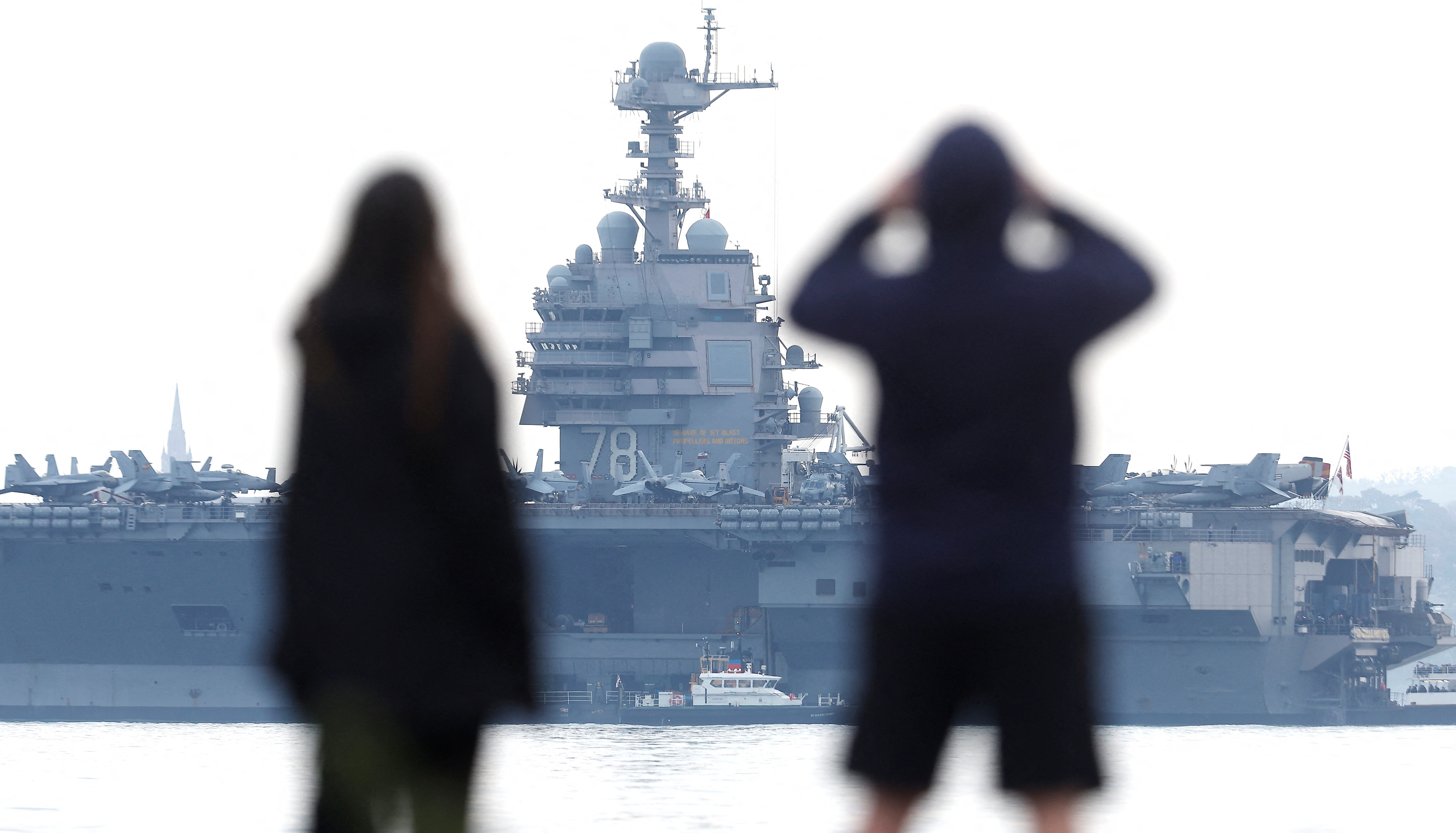 A couple stand on a beach near the USS Gerald R Ford aircraft carrier, currently anchored in the Solent near Gosport