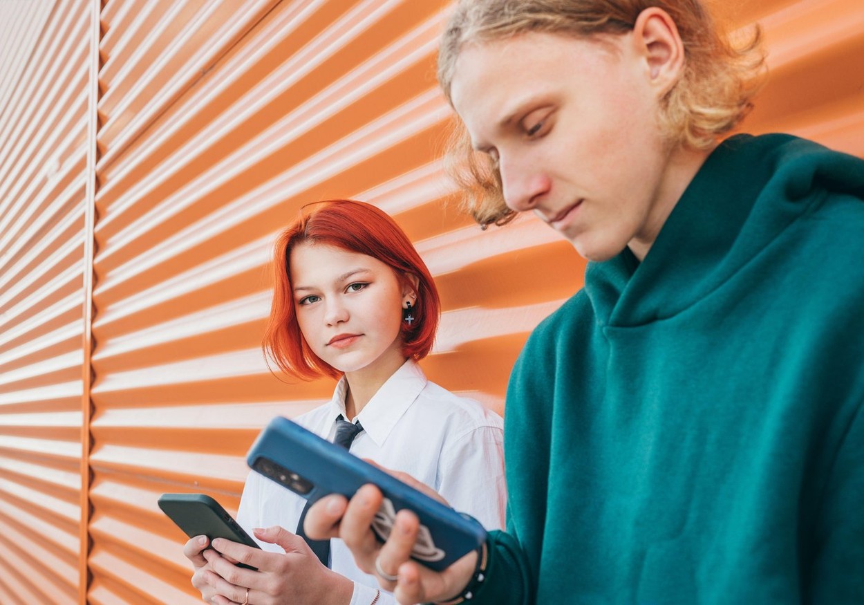Young caucasian teen girl portrait gazing at camera. She browsing smartphone devices with a friend boy standing near orange wall and. Careless young t