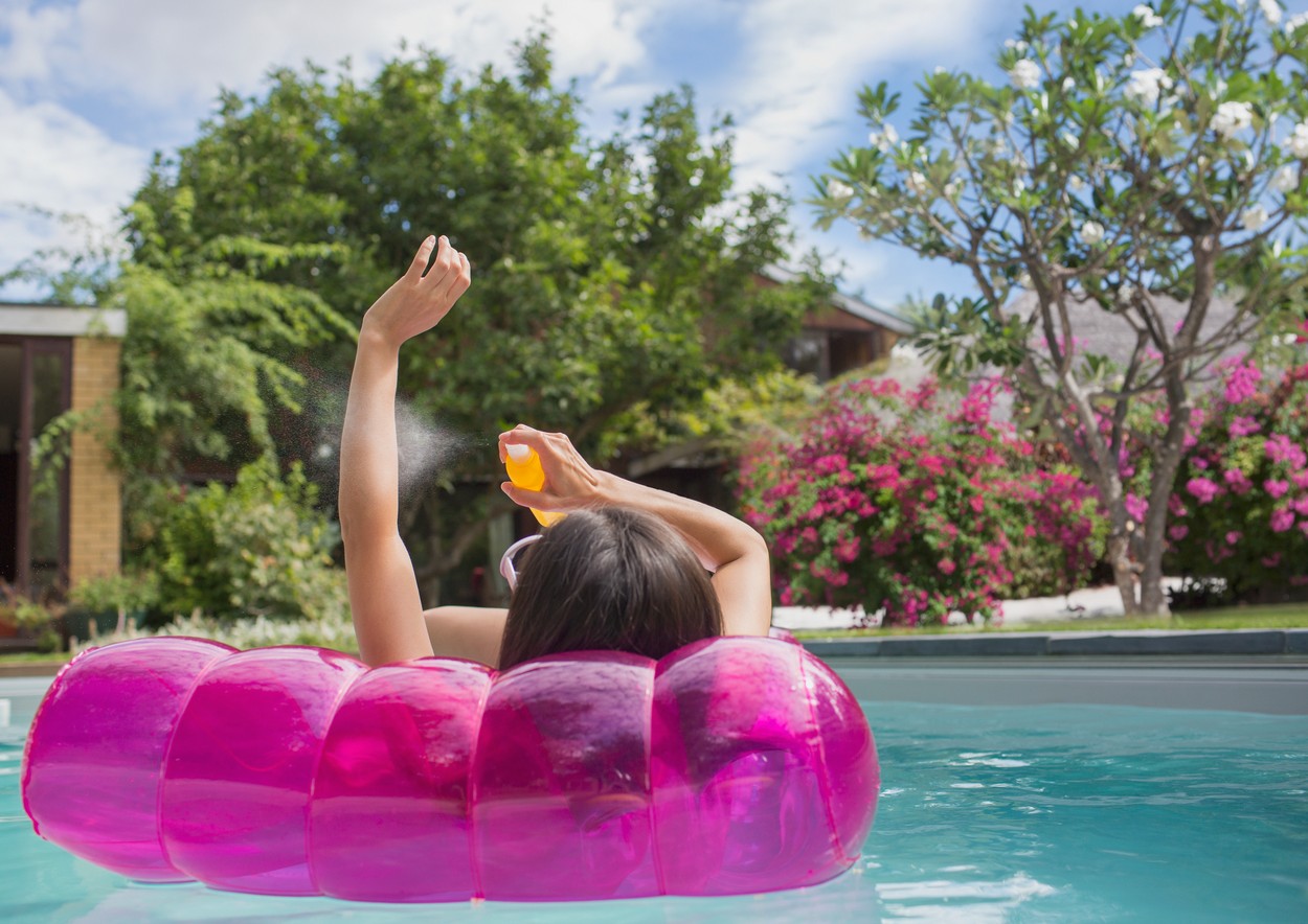 Woman applying, spraying sunscreen on arm on inflatable raft in sunny swimming pool