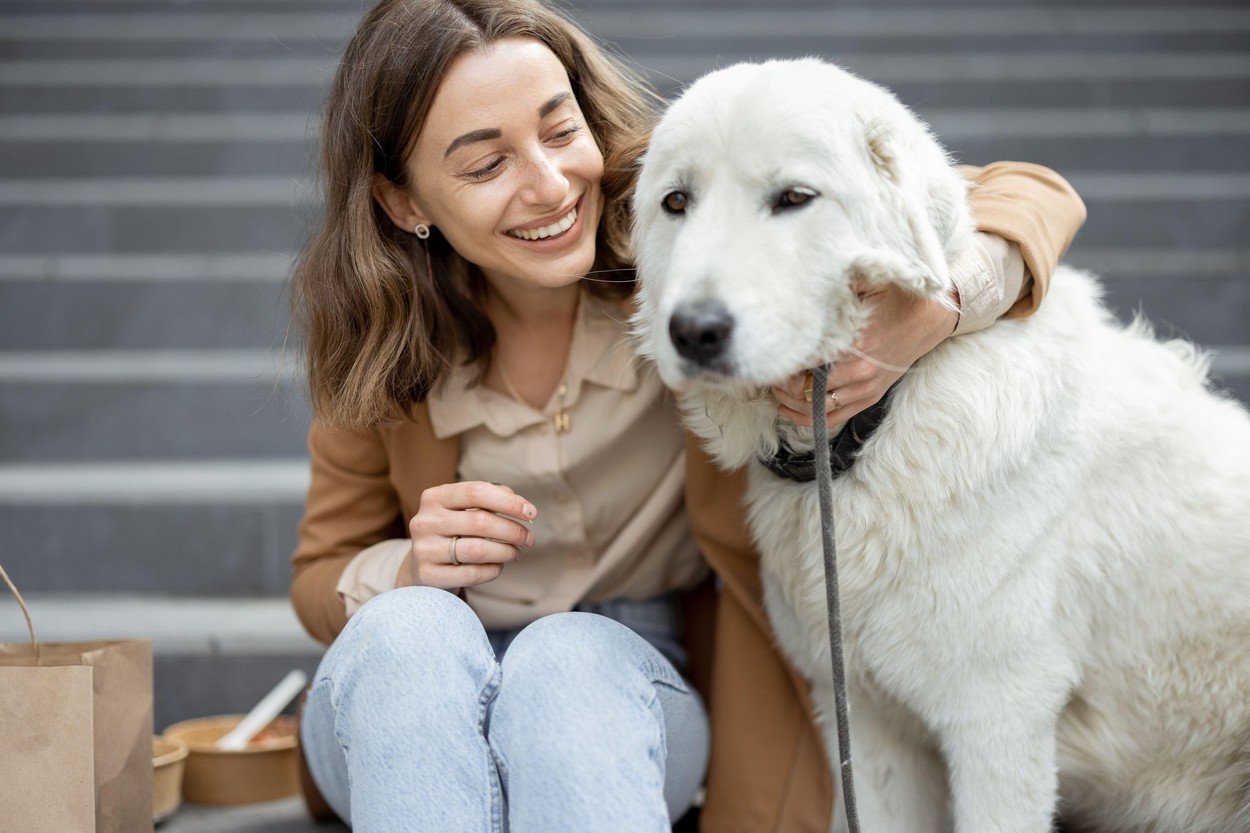 Pretty woman have outdoor lunch near office building with her big white dog while sitting on the stairs, hugs her dog. Pet friendly and pet care concept. Animal lover.
