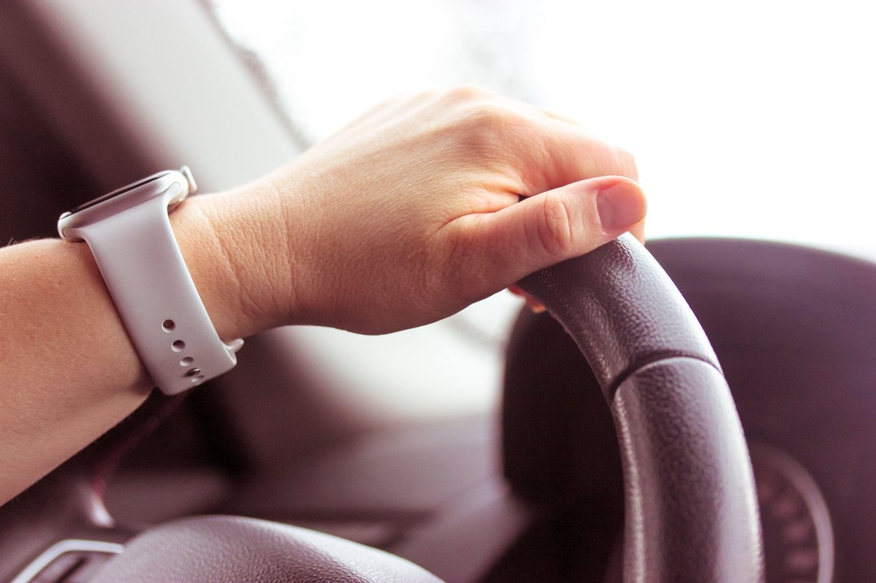 A woman's hand with a white watch is holding a black car steering wheel close-up. Traffic on a road. Driving lessons concept. Car interior, speedomete