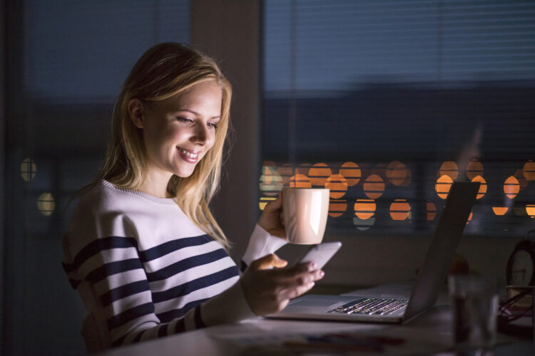Woman,At,Desk,,Holding,Smartphone,,Working,On,Laptop,At,Night.