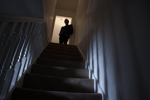 Silhouette of a man leaning against the wall at the top of a stairway, shadows cast on the walls from the light below.