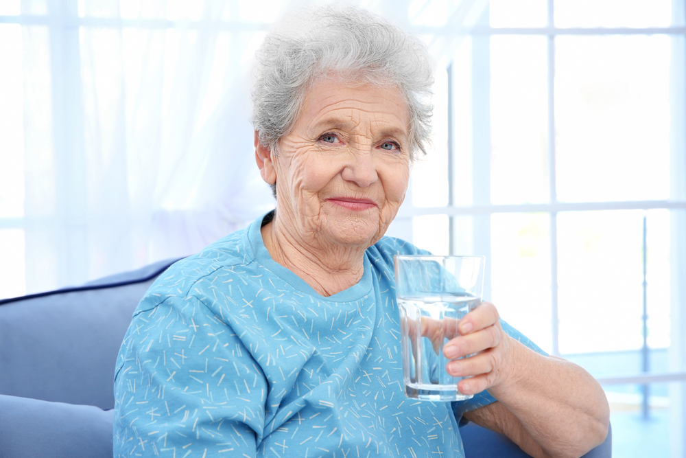 Elderly,Woman,Sitting,On,Couch,And,Holding,Glass,Of,Water.