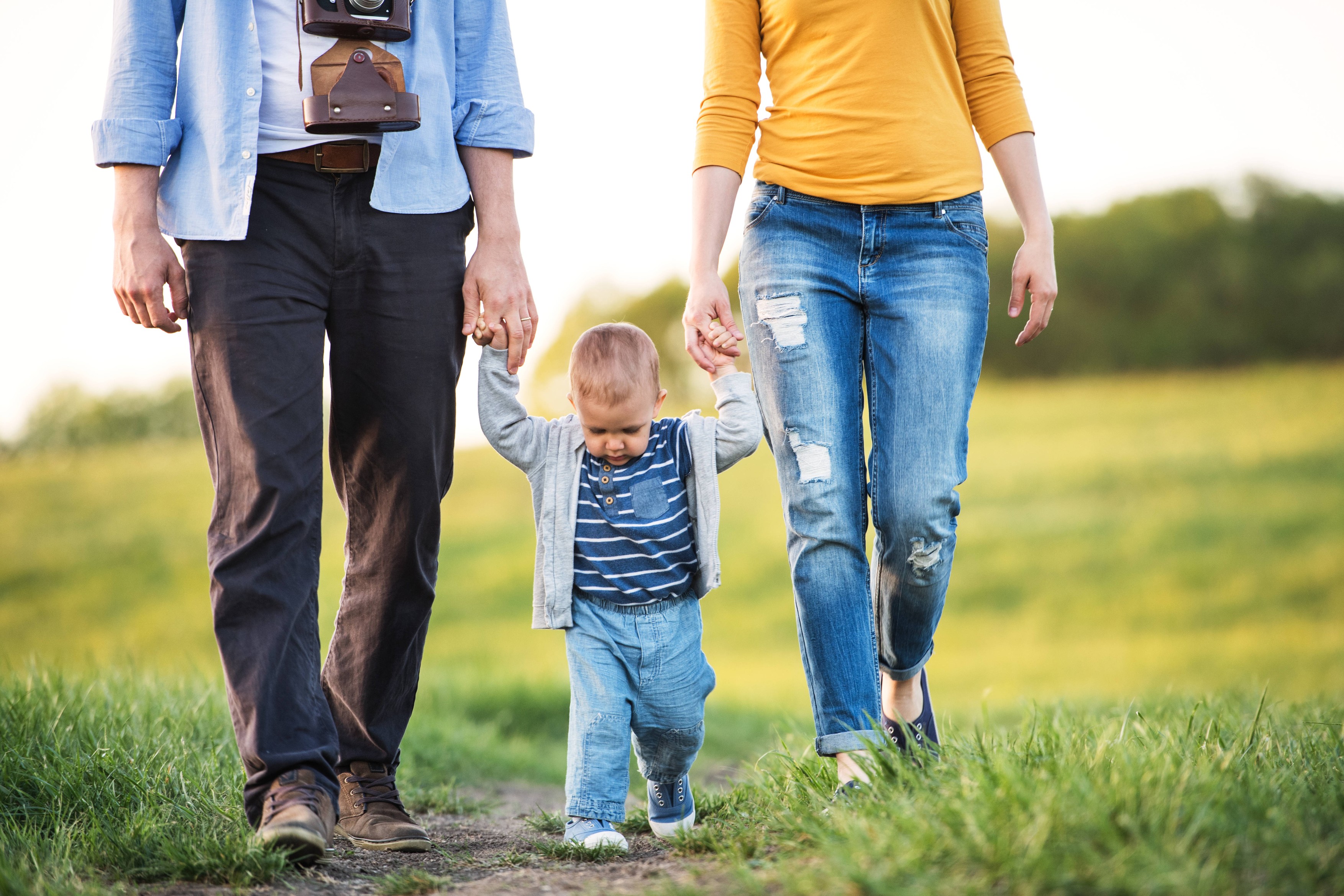 New parents holding small toddler, by hands, swinging him. Family outdoors on walk in spring nature. Happy family moment