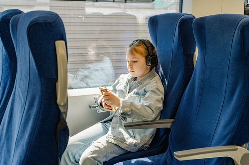 Little girl traveling on train in headphones looking at mobile phone. A little traveler on a train with blue chairs sits alone. Selected focus. High q