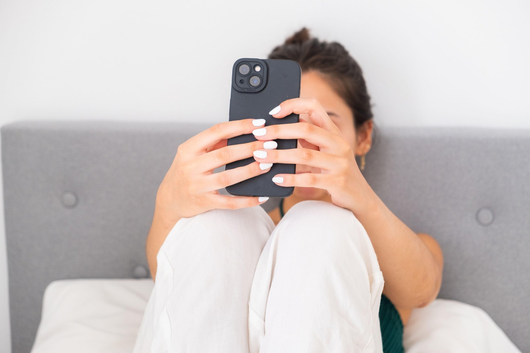 Close-up of a young teenager dressed in white linen pants sitting on her bed. She is taking a selfie for her social media and digital life with her sm