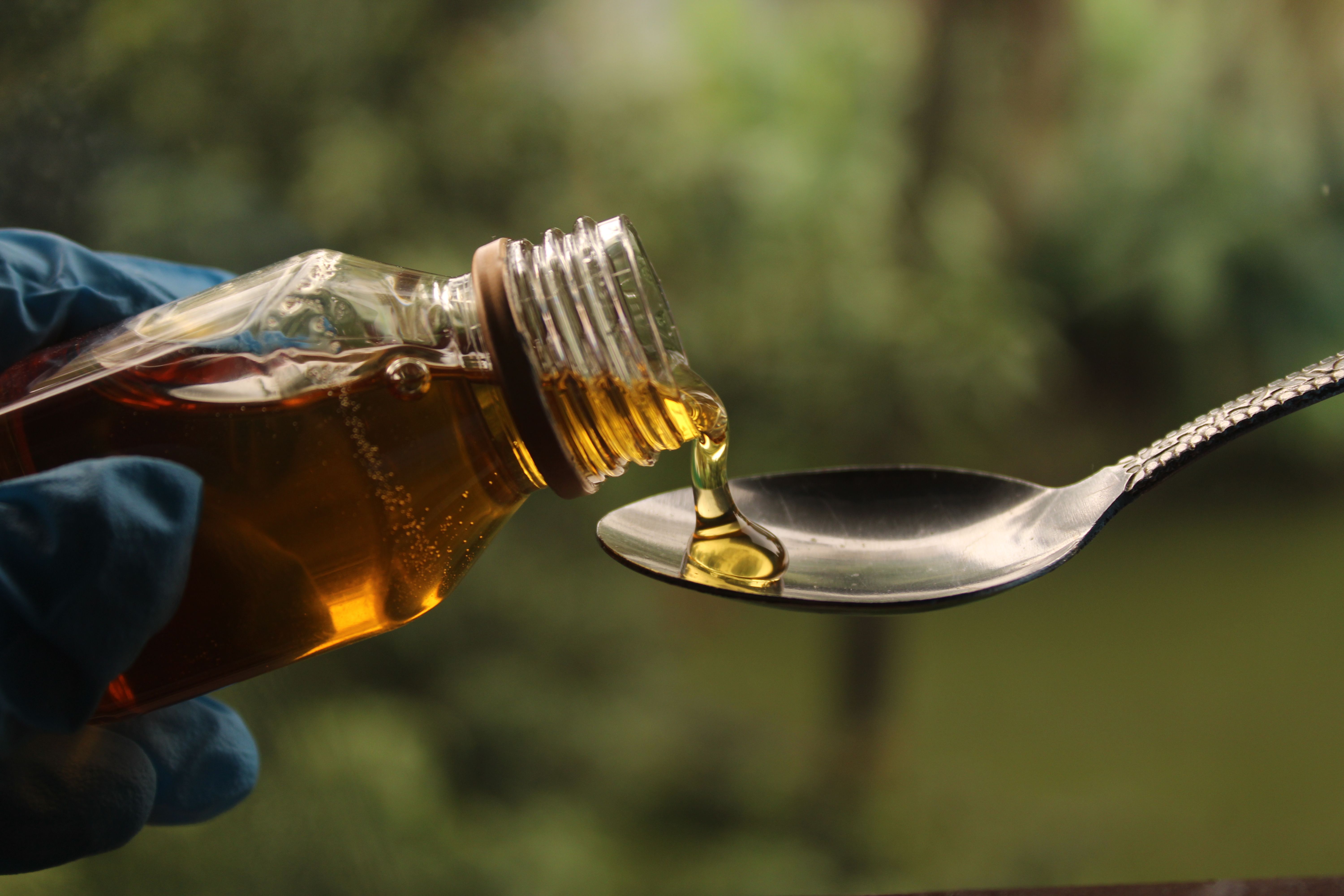 A gloved hand pours honey from a jar into a spoon against a blurred green background.