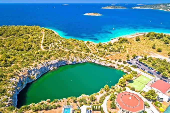 Aerial view of a coastal landscape showcasing a lush green area surrounding a circular lake, with the sea visible in the distance.
