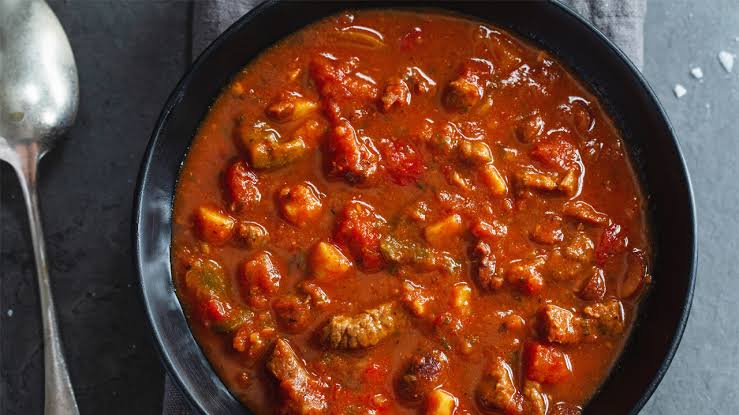 A bowl of hearty beef stew with chunks of meat and vegetables in a rich, savory broth, placed on a dark surface next to a spoon.