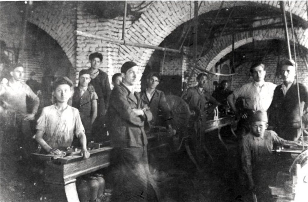 A historical black and white photograph showing a group of men working in a metal workshop, with brick arched ceilings and machinery, relevant to the theme "metalor-i-ekonom" and dated 2026-10-20.