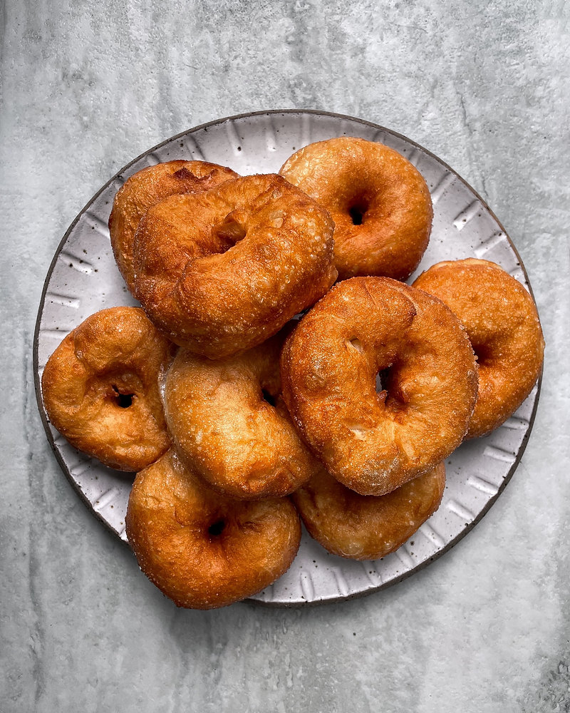 A plate of freshly made, golden-brown doughnuts arranged on a textured surface.