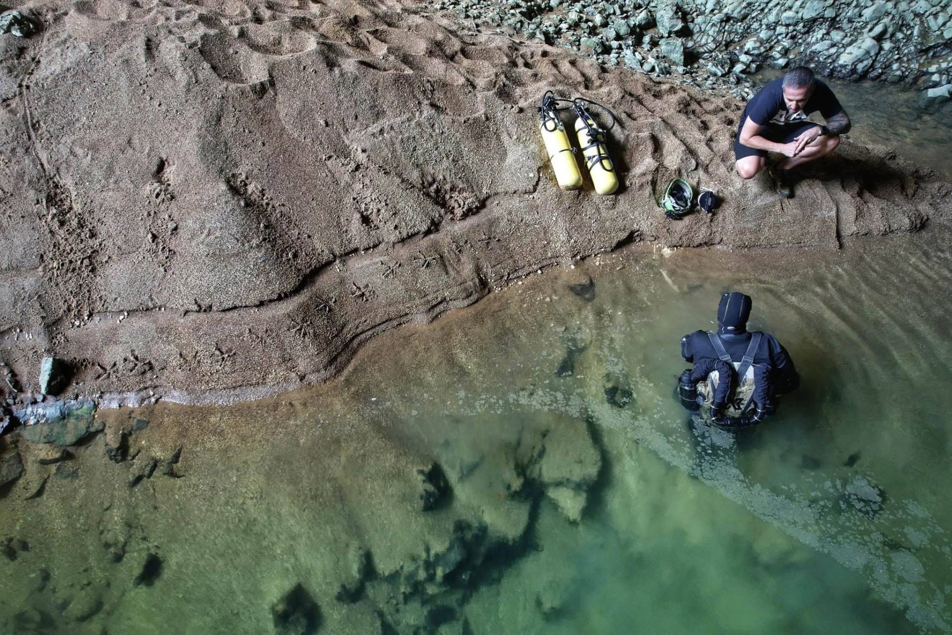 A person in a wetsuit prepares to dive into a cave water area with scuba gear while another person observes from the shore.