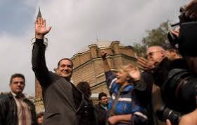 A person in a crowd waves enthusiastically at an outdoor event, with several attendees and a historic building in the background.
