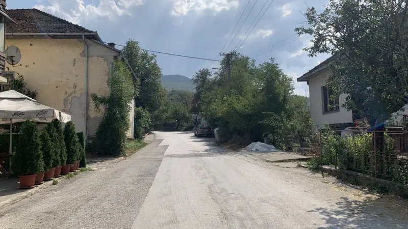 A quiet rural street scene with houses on either side, lined with trees and bushes under a partly cloudy sky. The road is unpaved and stretches into the distance, suggesting a tranquil countryside setting.