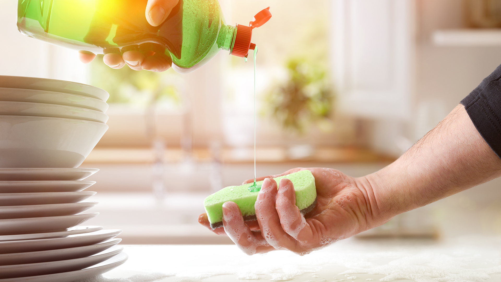 A person pouring dish soap onto a sponge in a bright kitchen, with sunlight filtering through a window and a stack of clean dishes beside them.