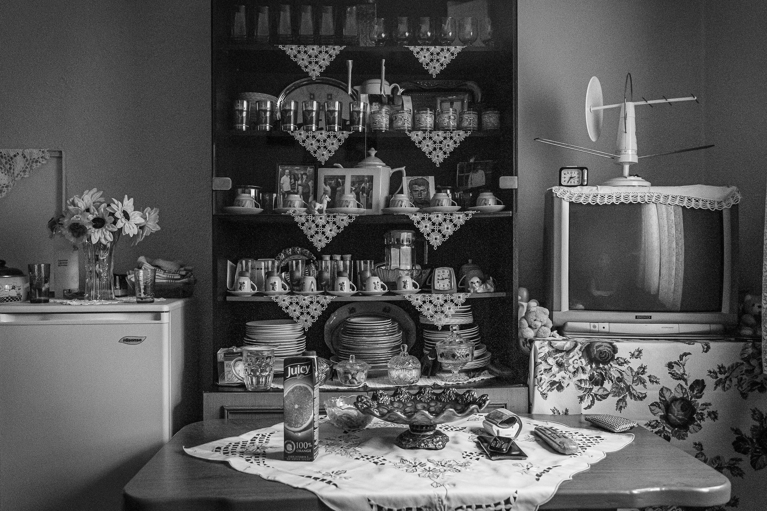Fotografija iz ciklusa "Potočarske sjene" atuora Velije Hasanbegovića prikazuje unutrašnjost kuće jedne od majki u Potočarima, sa vitrinom ispunjenom posuđem i porodičnim fotografijama, stolom i starim televizorom sa antenom.
