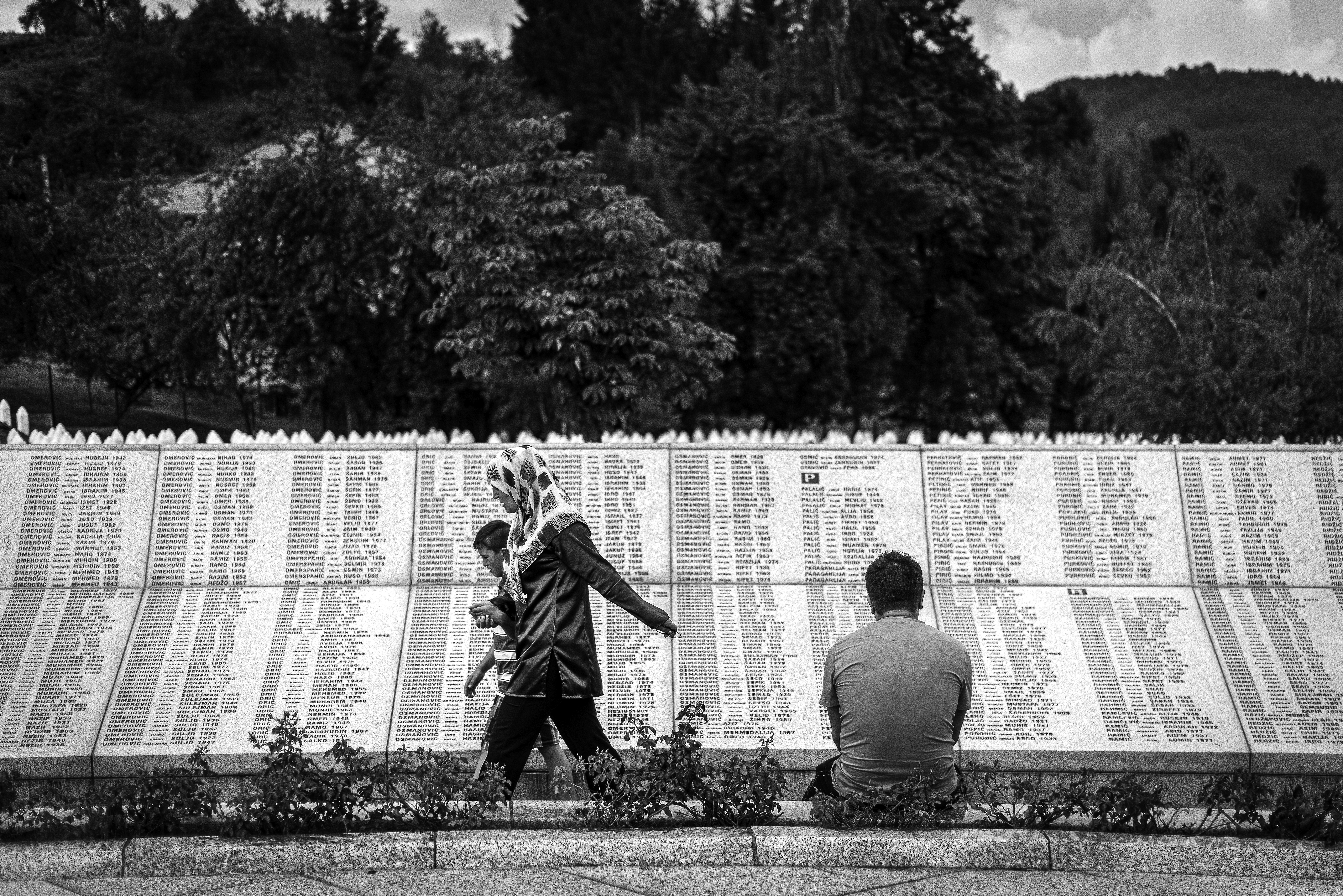 Fotografija iz ciklusa "Potočarske sjene" autora Velije Hasanbegovića prikazuje posjetitelje ispred zida sa imenima žrtava u Memorijalnom centru Srebrenica-Potočari.