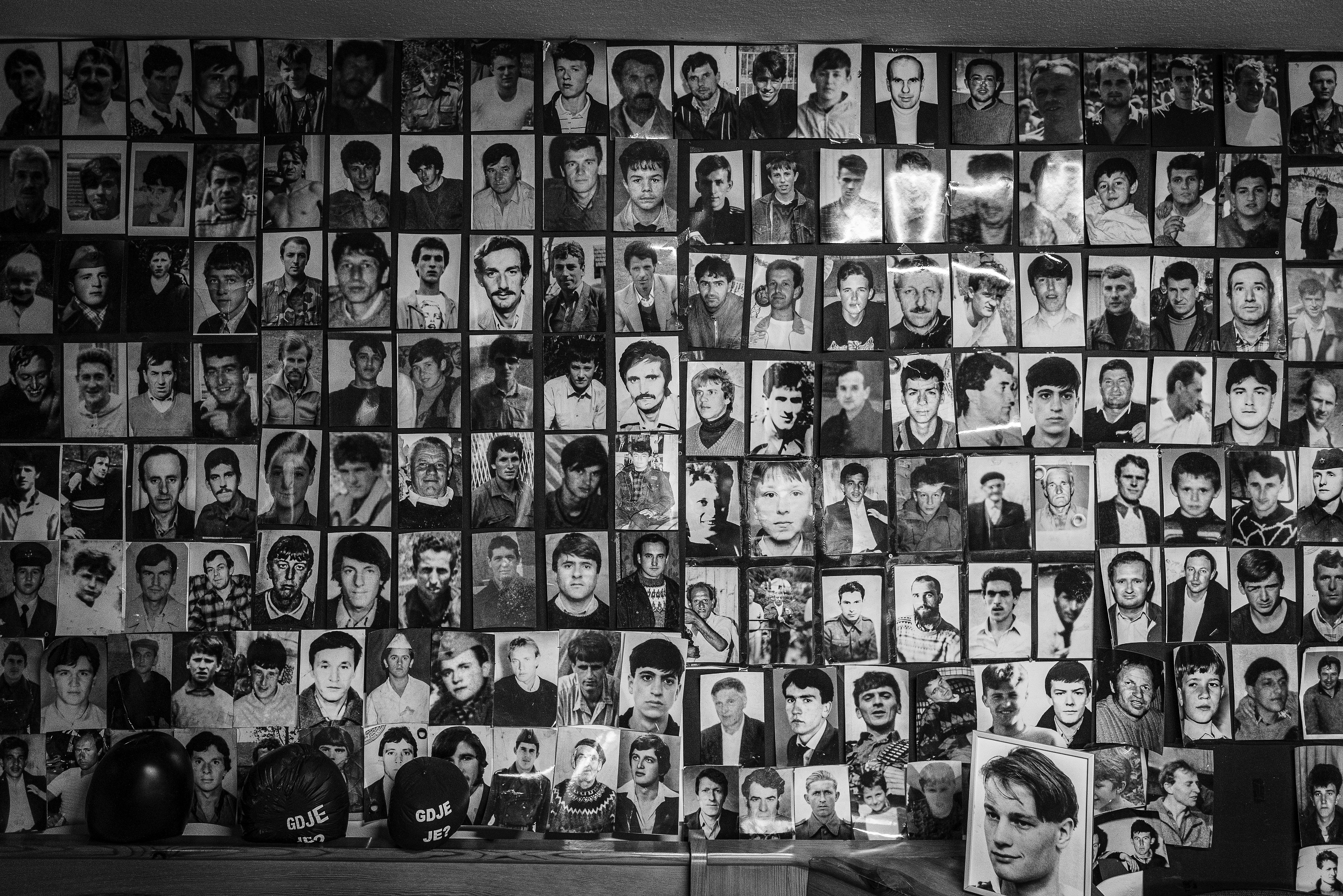 Fotografija iz ciklusa "Potočarske sjene" Velije Hasanbegovića prikazuje zid ispunjen portretima žrtava genocida u Srebrenici i ličnim uspomenama koje čuvaju sjećanje na ubijene.