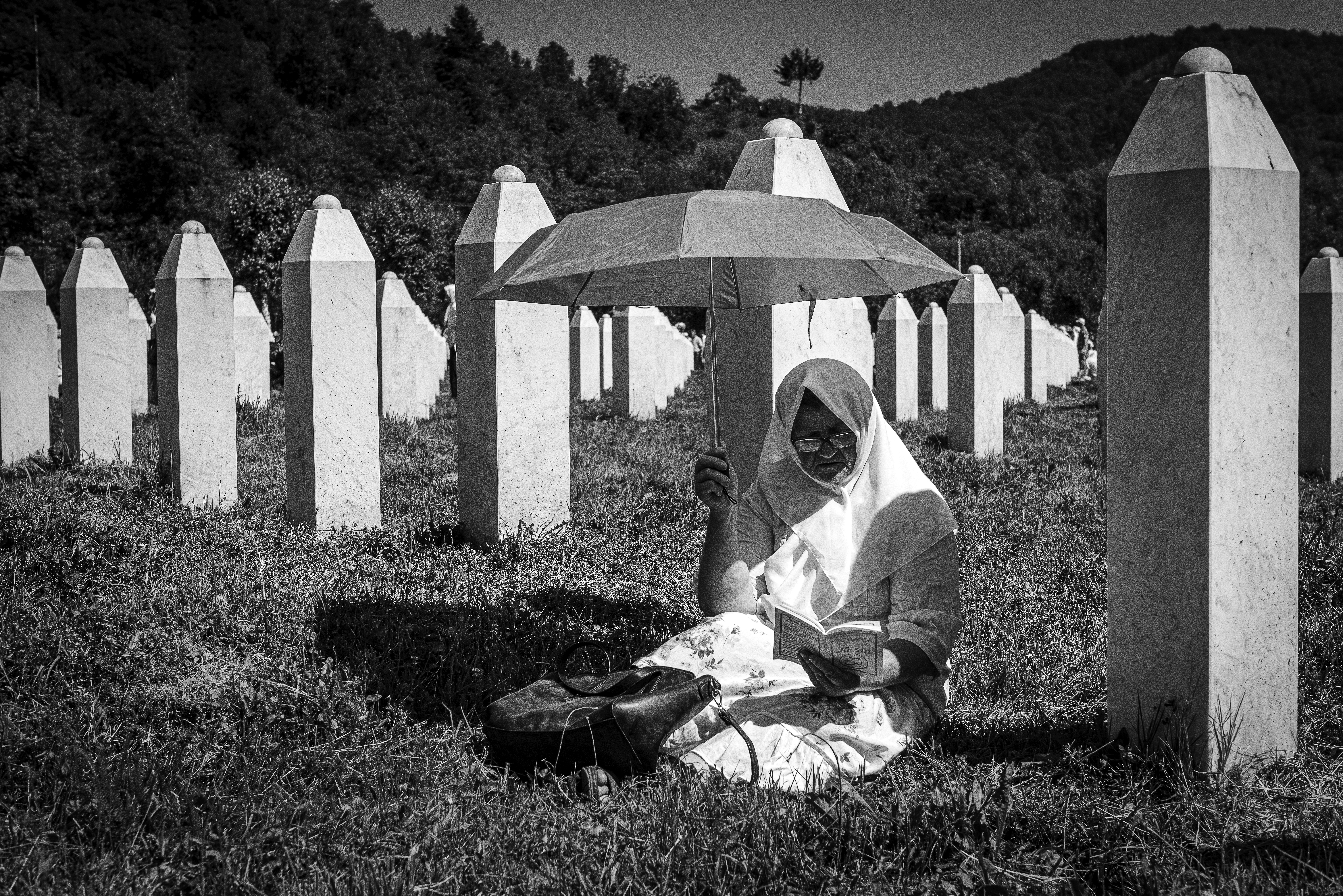 Fotografija iz ciklusa "Potočarske sjene" Velije Hasanbegovića prikazuje ženu sa kišobranom koja uči dovu među mezarima u Memorijalnom centru Srebrenica-Potočari.